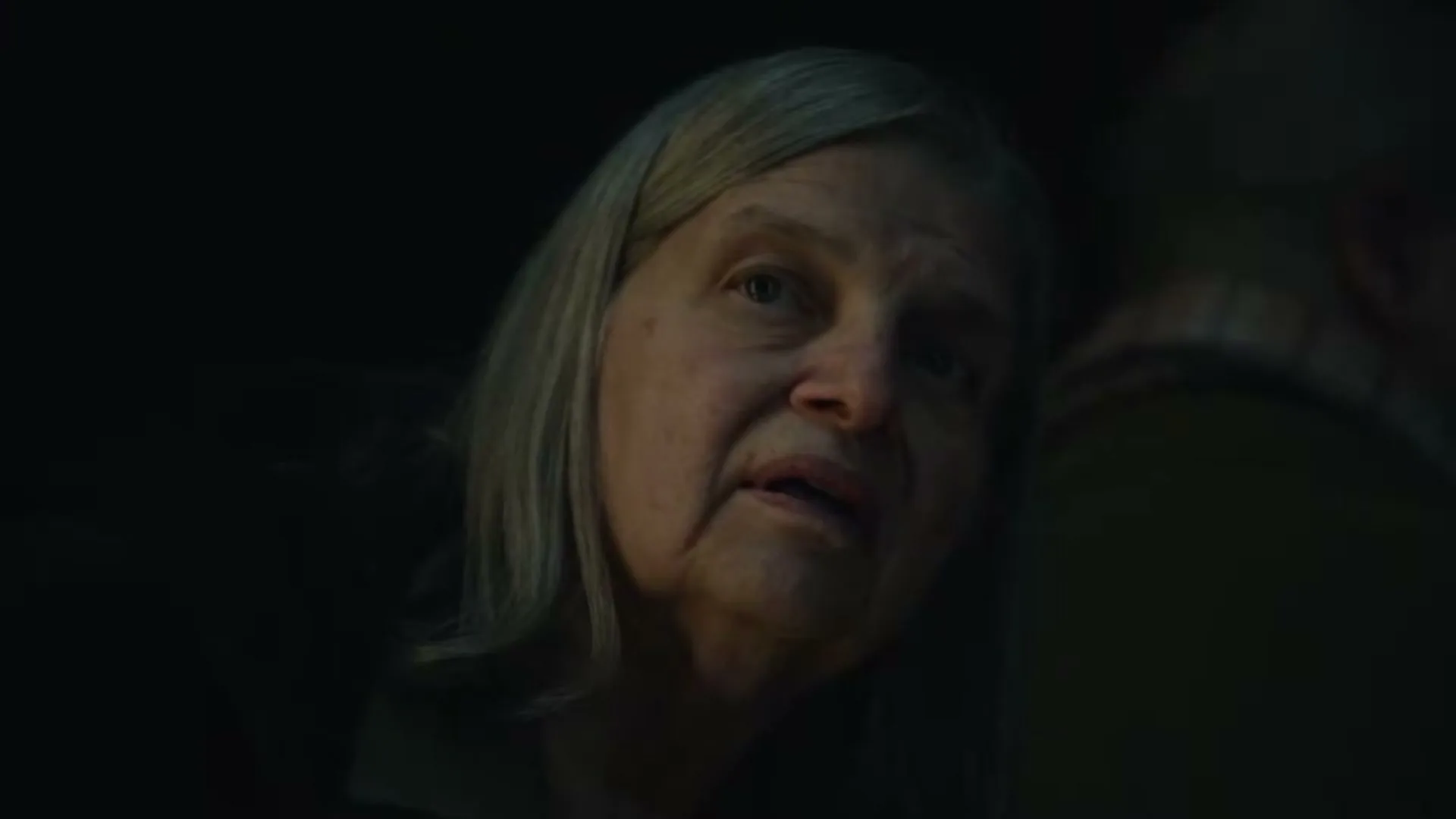 An older woman with gray hair looks up with a surprised expression in a dimly lit interior setting.