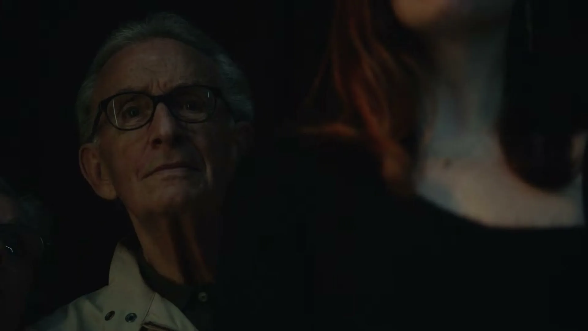 A senior man with glasses and a light jacket looks up intently, partially obscured by a woman's shoulder in a dark, crowded room.