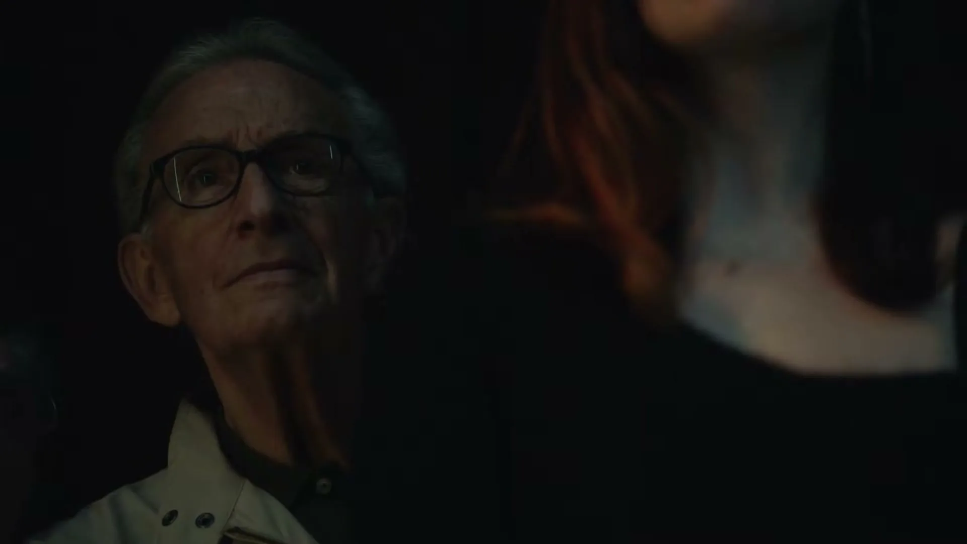 An older man with glasses and a light jacket looks up intently, partially obscured by a woman's dark shoulder in a dimly lit interior.