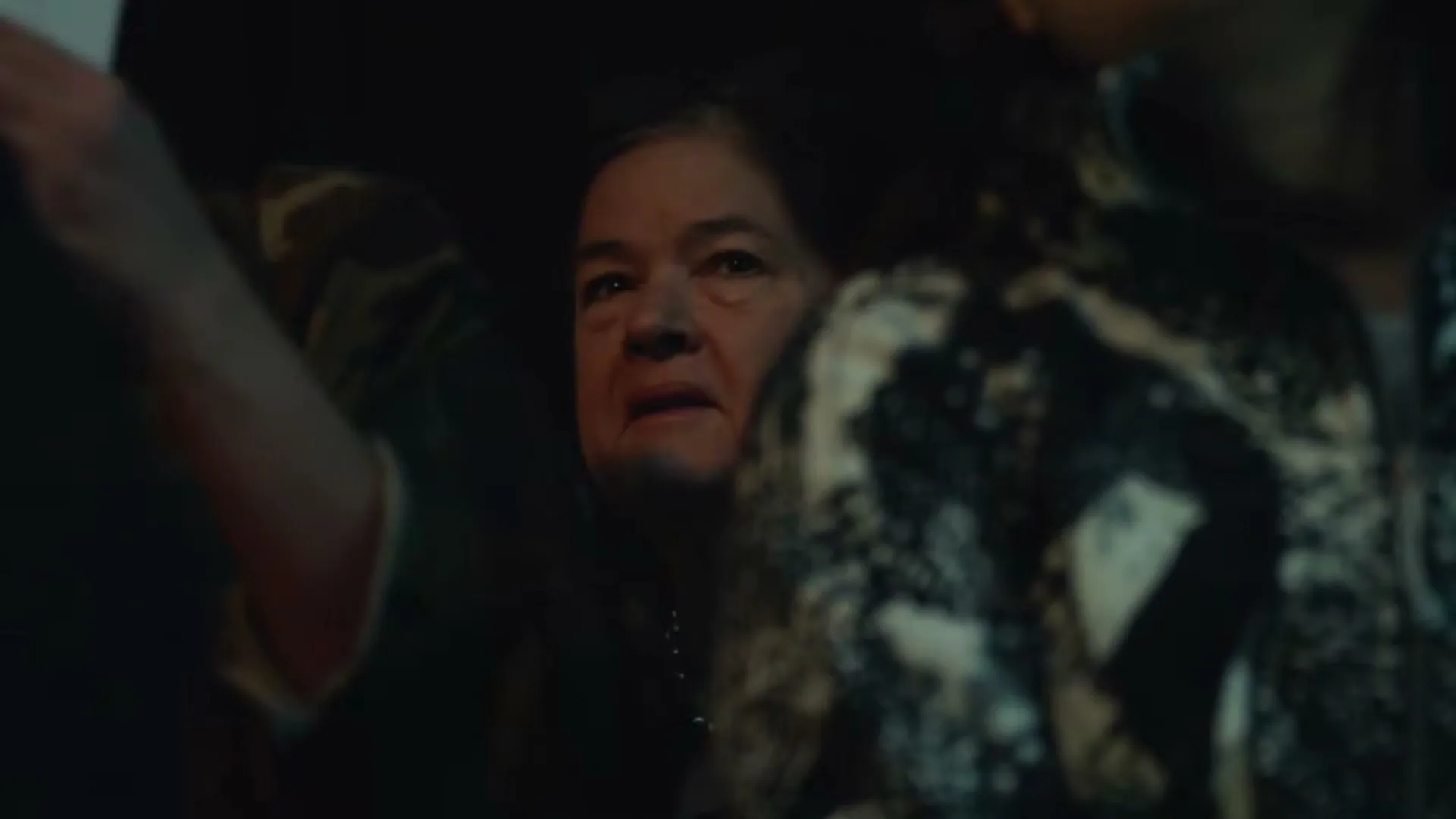 A woman in her 60s with dark hair and a pearl necklace looks on with a concerned expression, surrounded by other people in a dark, crowded interior.