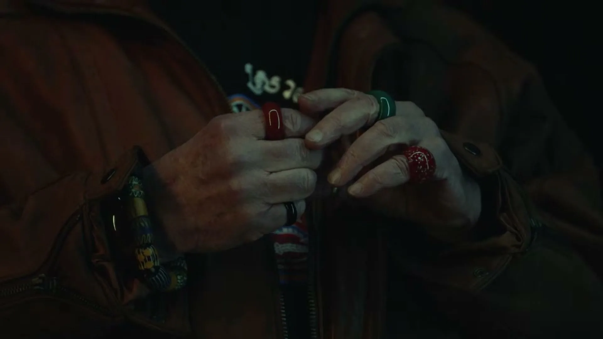A person's hands, adorned with colorful rings and bracelets, are clasped together in a dimly lit interior setting.