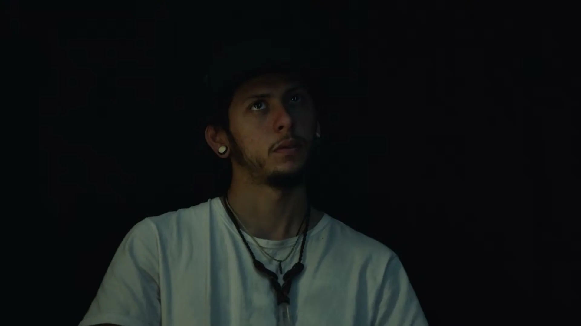 A young man with a beard and a white t-shirt looks up with a curious expression in a dimly lit, dark room.