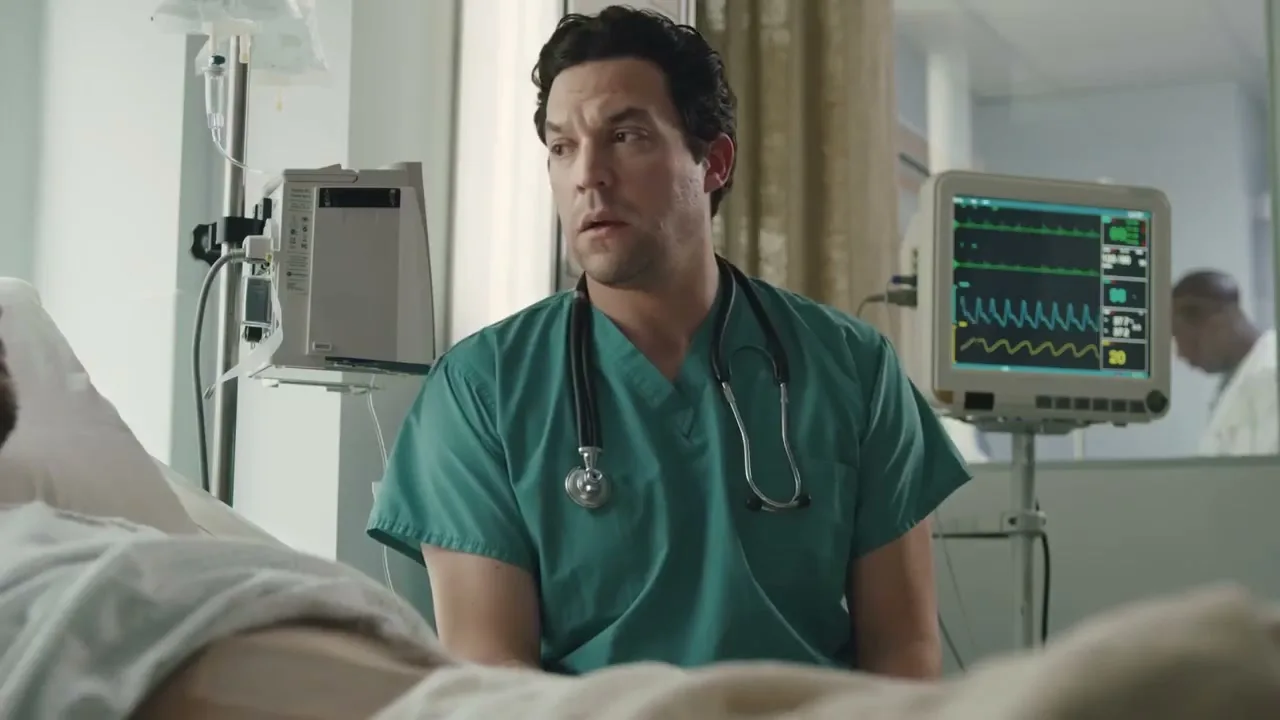 A male doctor in green scrubs and a stethoscope around his neck sits by a patient's bed in a hospital room, looking concerned.