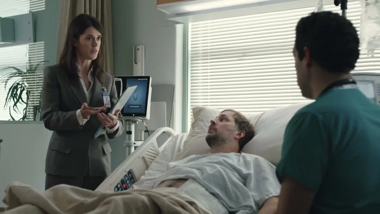 A woman in a suit talks to a man lying in a hospital bed, while a male nurse stands nearby in a bright hospital room.