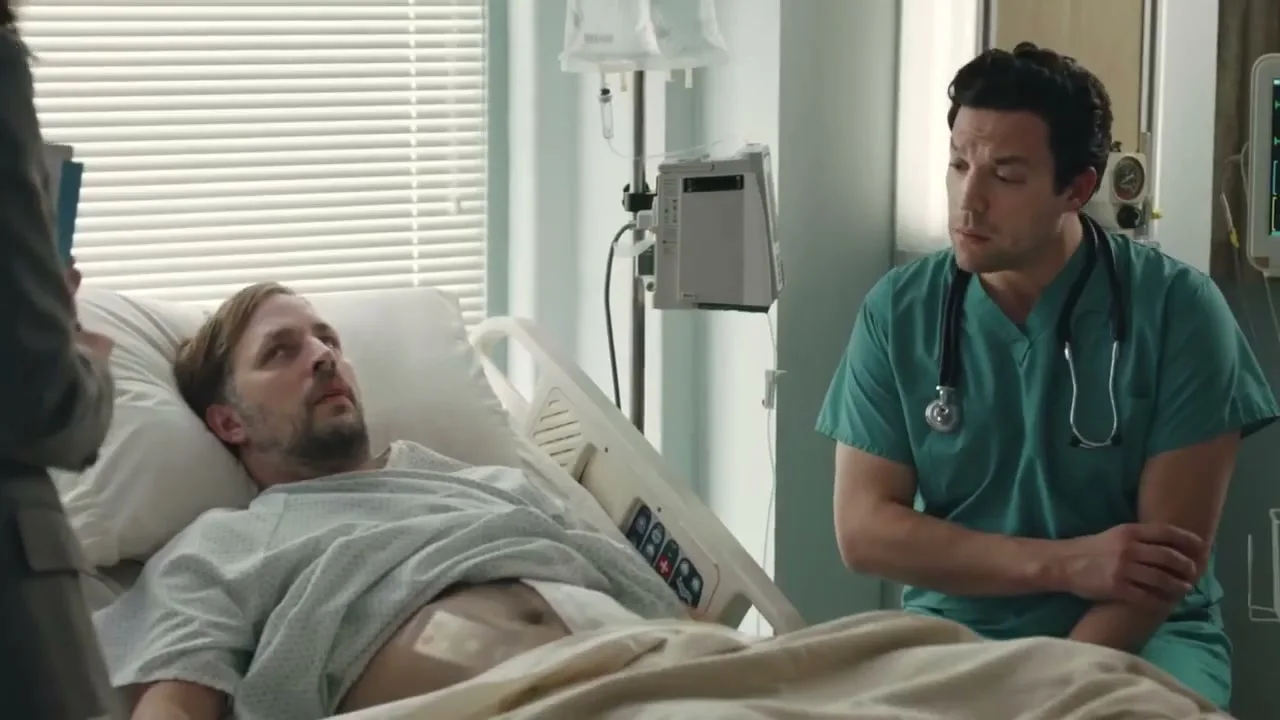 A male patient lies in a hospital bed, looking up at a male doctor in scrubs who is talking to him in a hospital room.