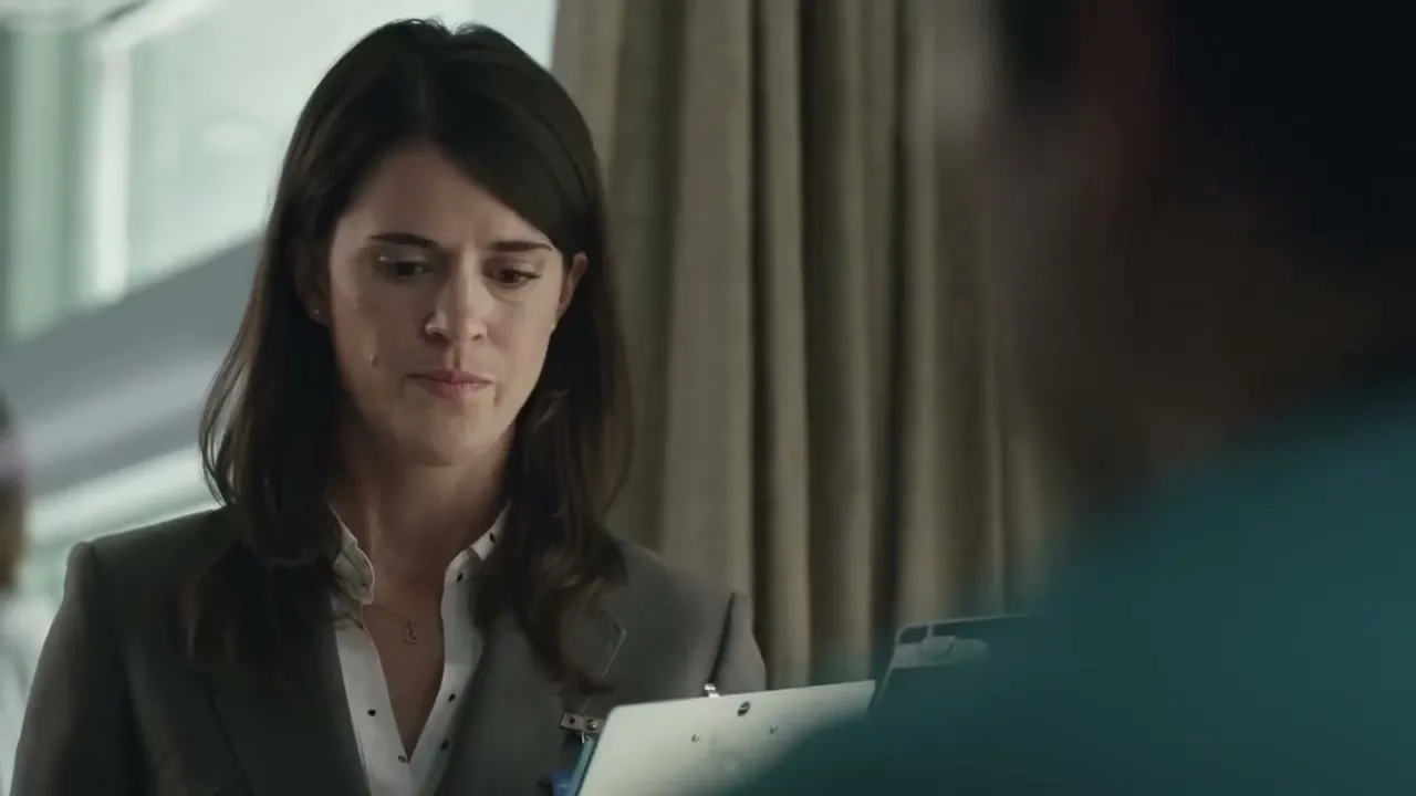 A female doctor in a grey suit and white shirt looks at a patient in a hospital room.