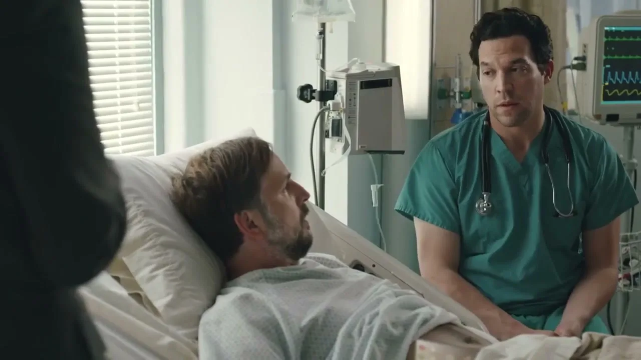 A male patient with a beard lies in a hospital bed, looking at a doctor in green scrubs who is sitting beside him in a hospital room.