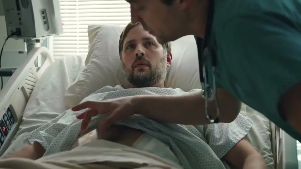 A male patient with a beard lies in a hospital bed, looking up at a male doctor in scrubs who leans over him, holding a stethoscope to his stomach.
