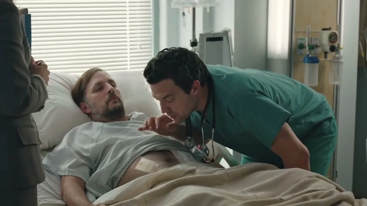 A male patient in a hospital gown lies in bed while a male doctor in scrubs leans over him, examining his stomach in a hospital room.