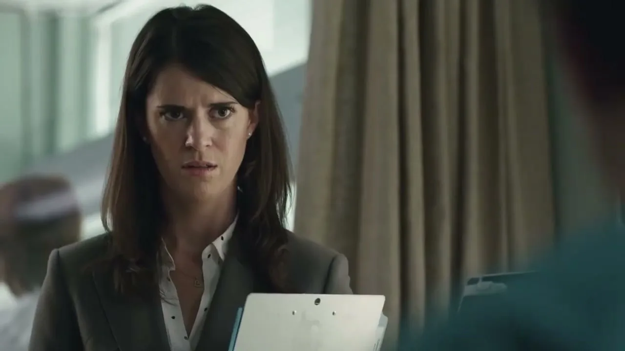 A female doctor in a grey suit looks confused and concerned while holding a clipboard in a hospital room.