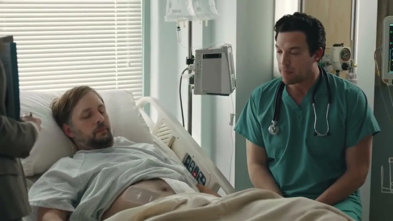 A male patient lies in a hospital bed, looking up at a male doctor in scrubs who is sitting beside him, while a female in a suit stands partially visible on the left.