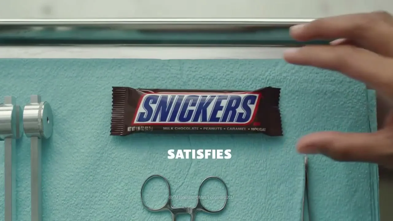 A hand places a Snickers bar on a surgical tray with medical instruments, and the word "SATISFIES" appears on screen.