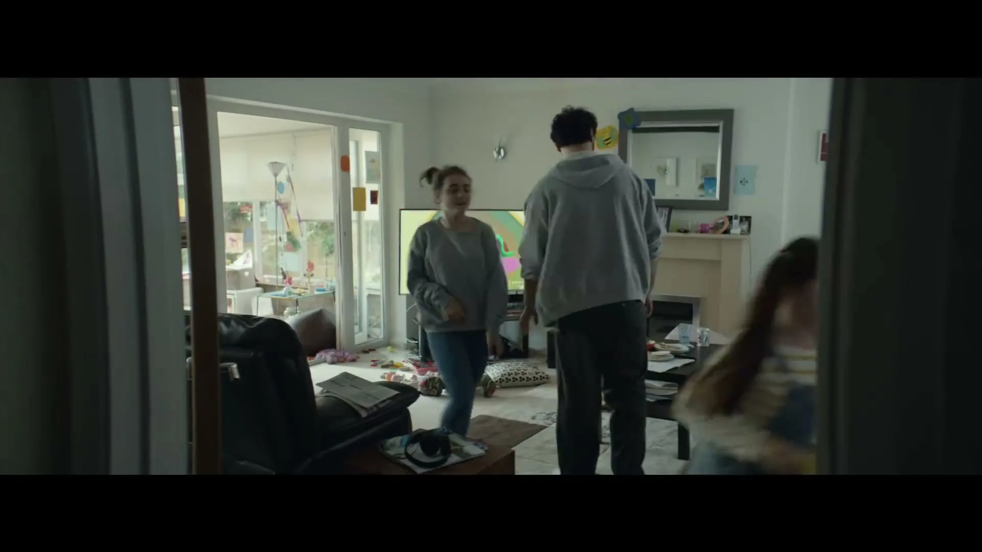 A family of three, a father and two daughters, are in a messy living room, with the father bending down to pick up something.