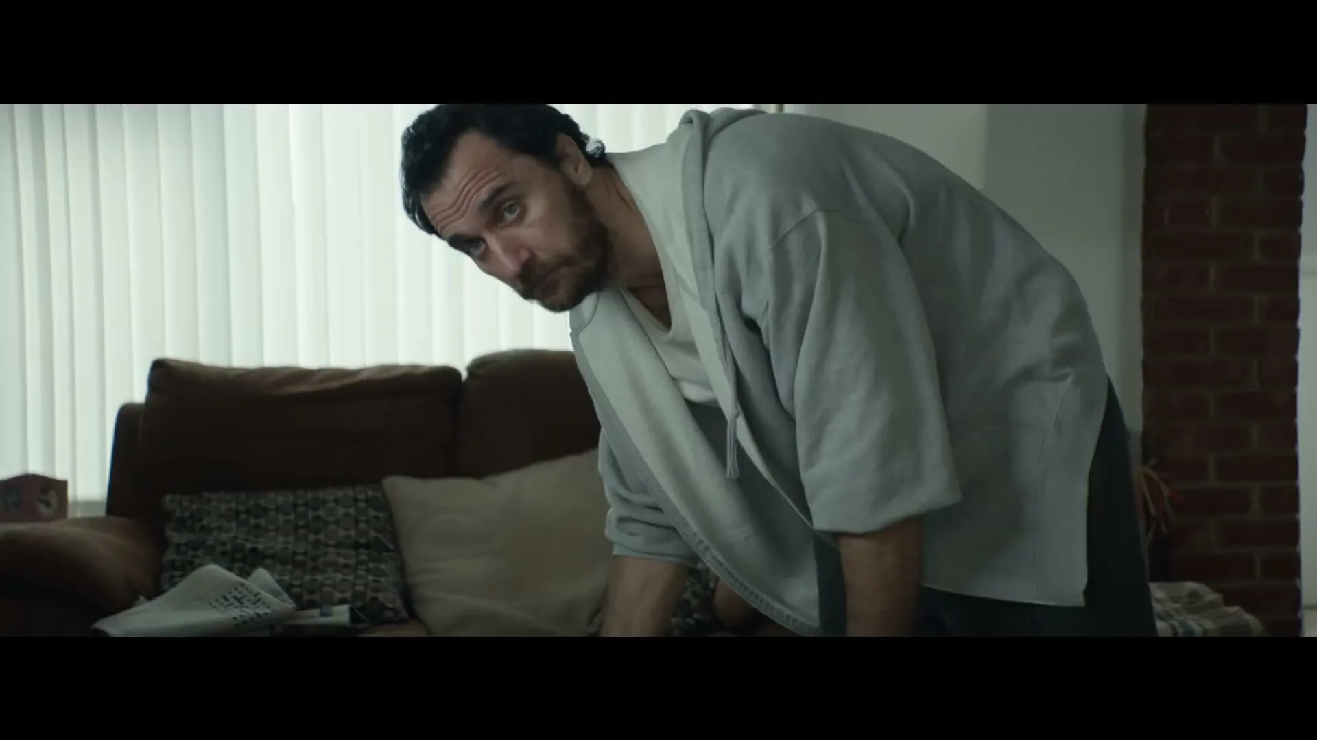 A man in a robe bends over a table with food, looking up with a concerned expression in a living room.