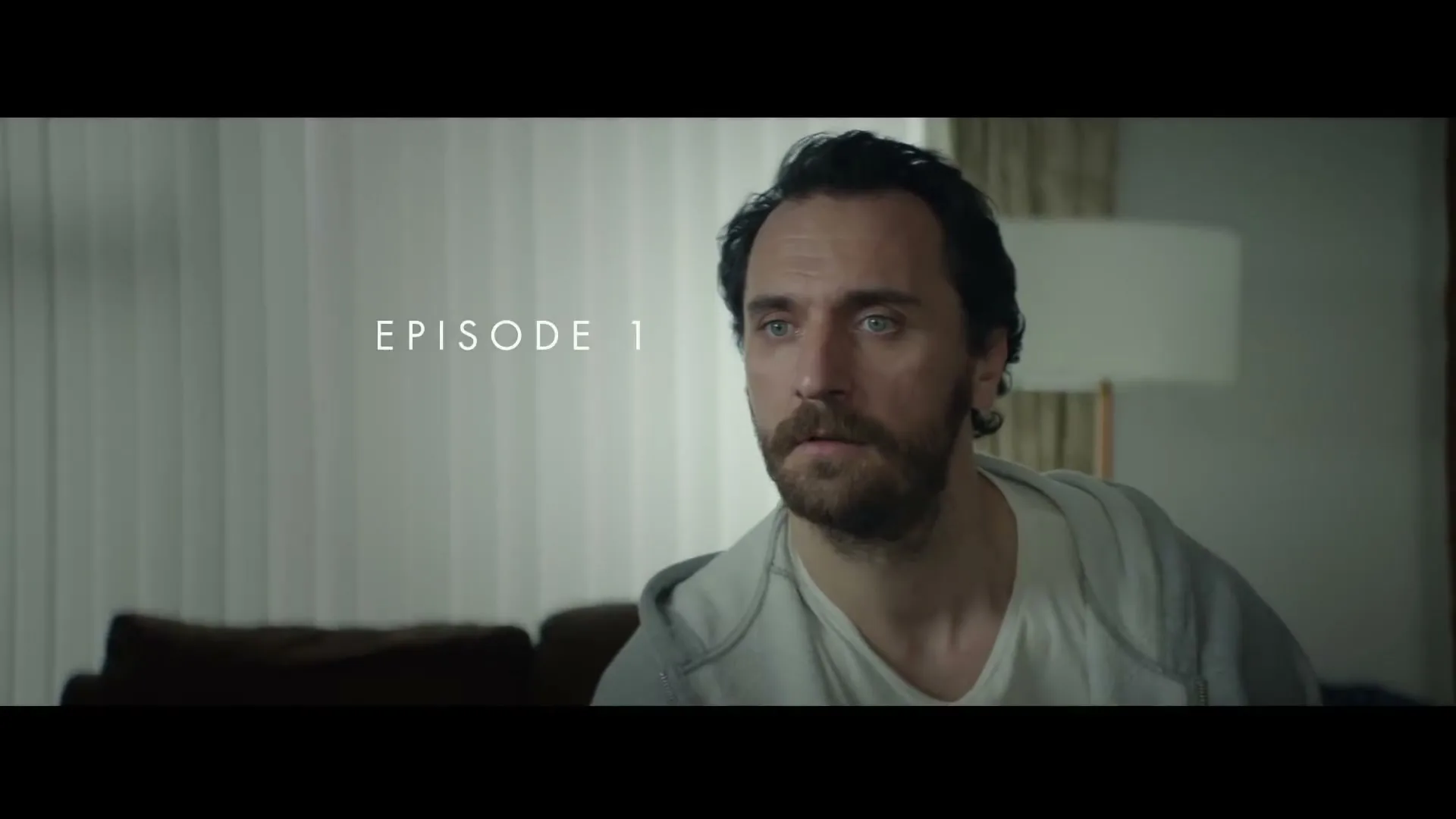 A man with a beard and dark hair looks around with a worried expression in a living room with vertical blinds.