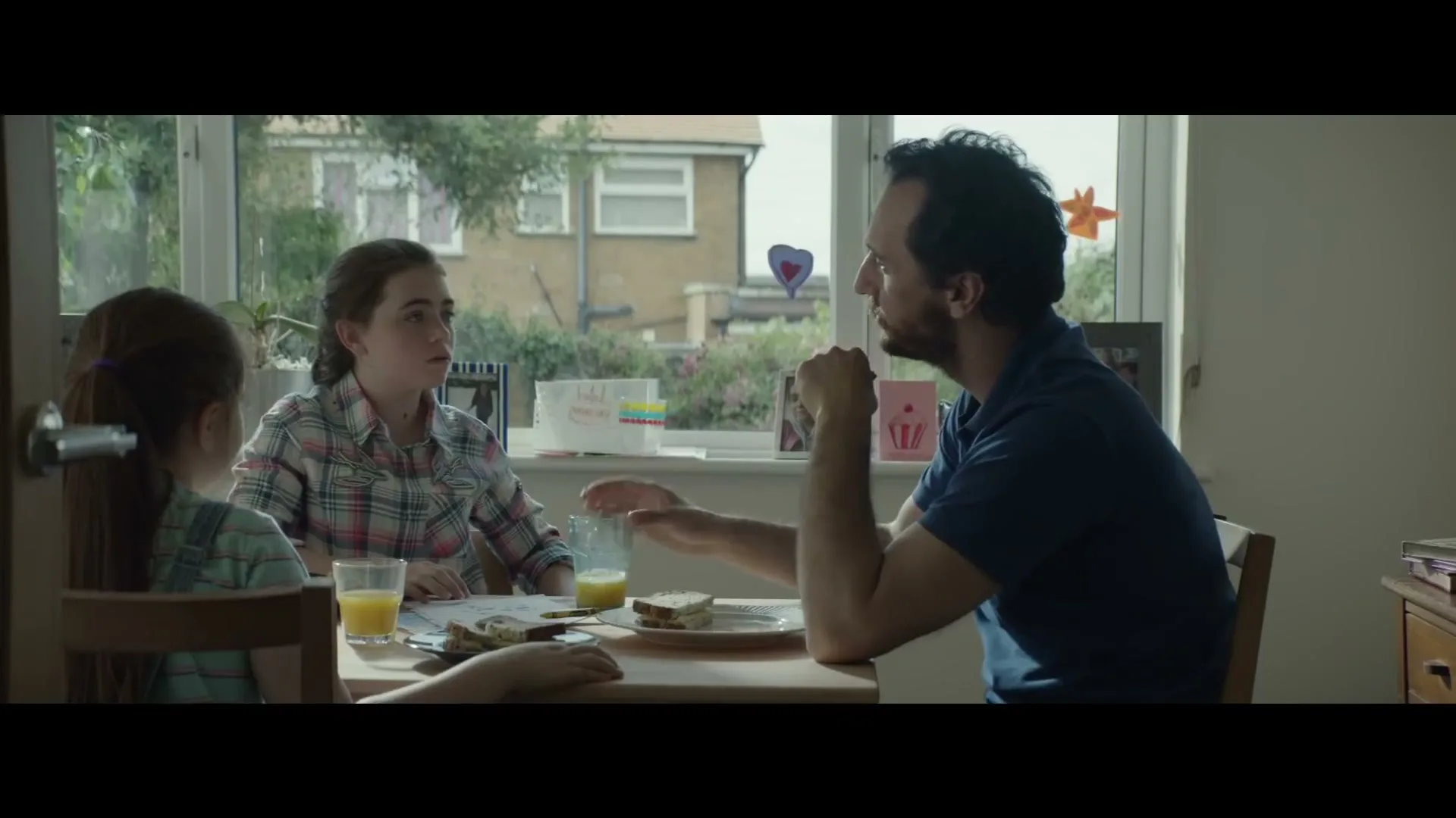 A father and his two daughters sit at a kitchen table, eating breakfast and talking, with a window showing houses in the background.