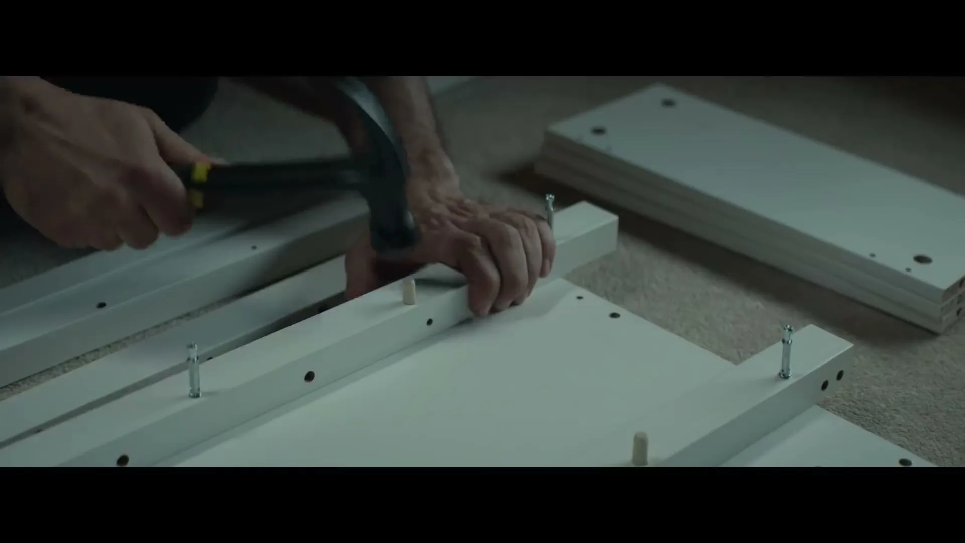 A man's hands are shown assembling white furniture pieces on a carpeted floor, using a hammer to tap in a wooden dowel.