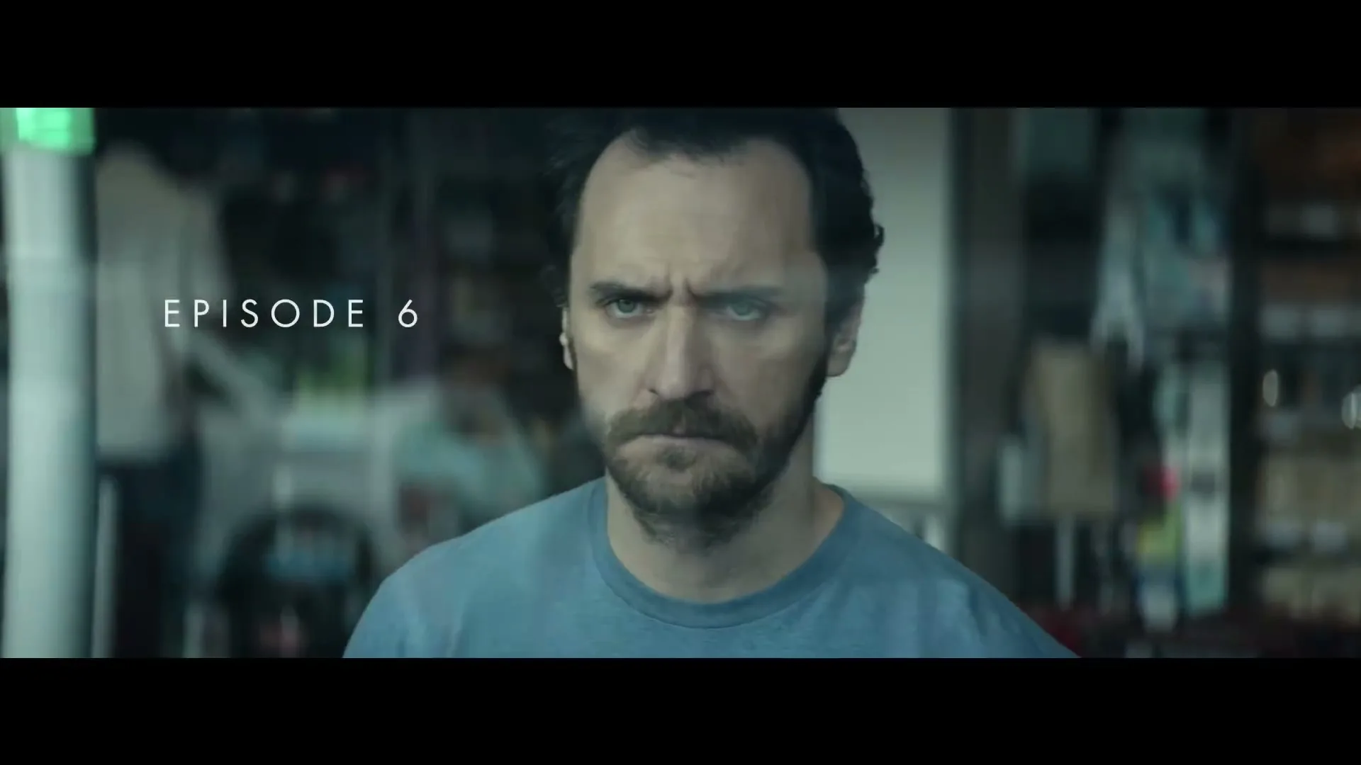 A man with a beard and dark hair looks intensely at the camera, with a blurred background of a store interior.