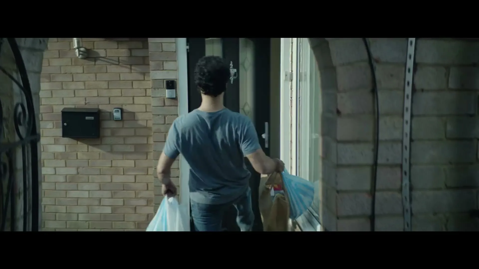 A man in a blue t-shirt and jeans carries grocery bags towards the front door of a brick house during the day.