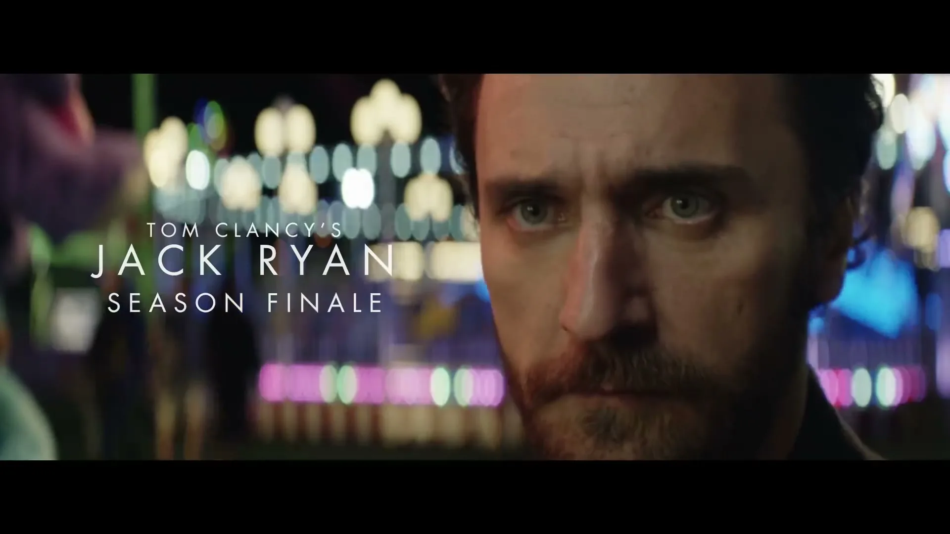 A man with a beard and intense eyes looks directly at the camera, with blurred amusement park lights in the background at night.