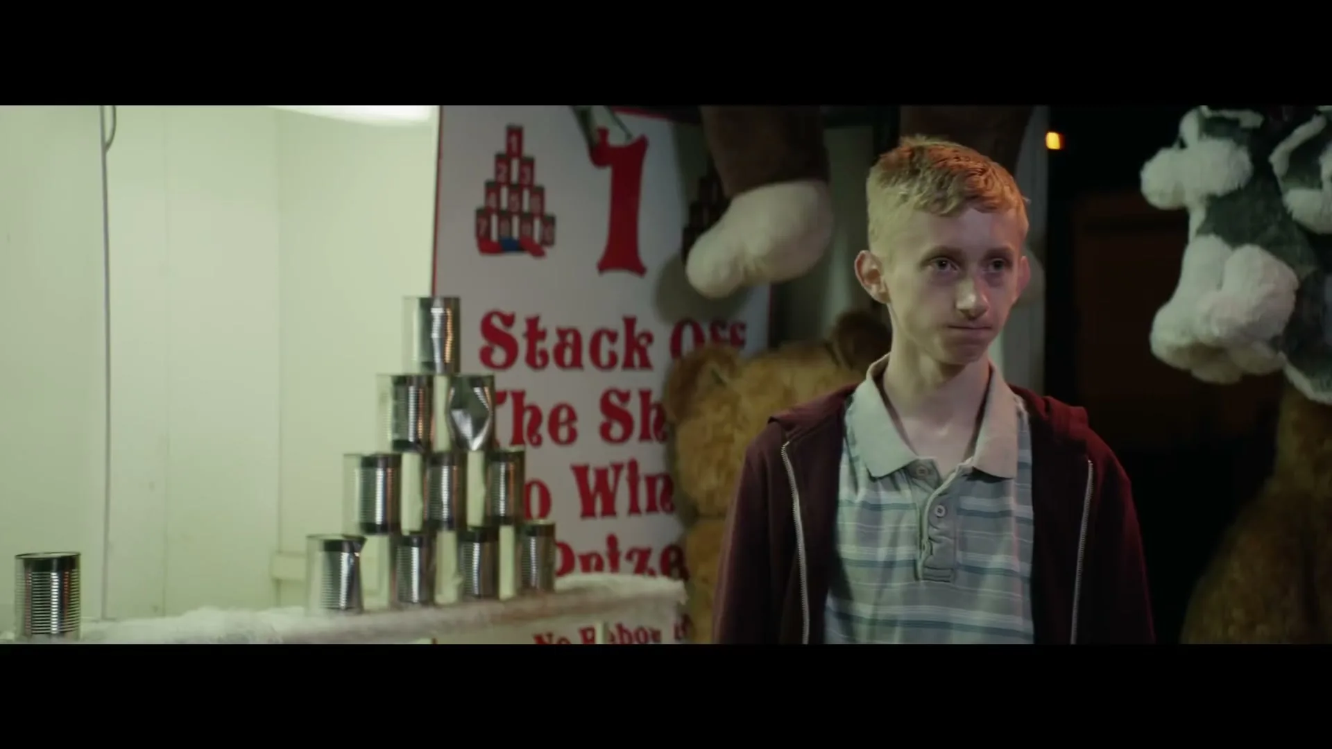 A young blonde male stands in front of a carnival game with stacked cans and stuffed animals, looking around with a neutral expression at night.
