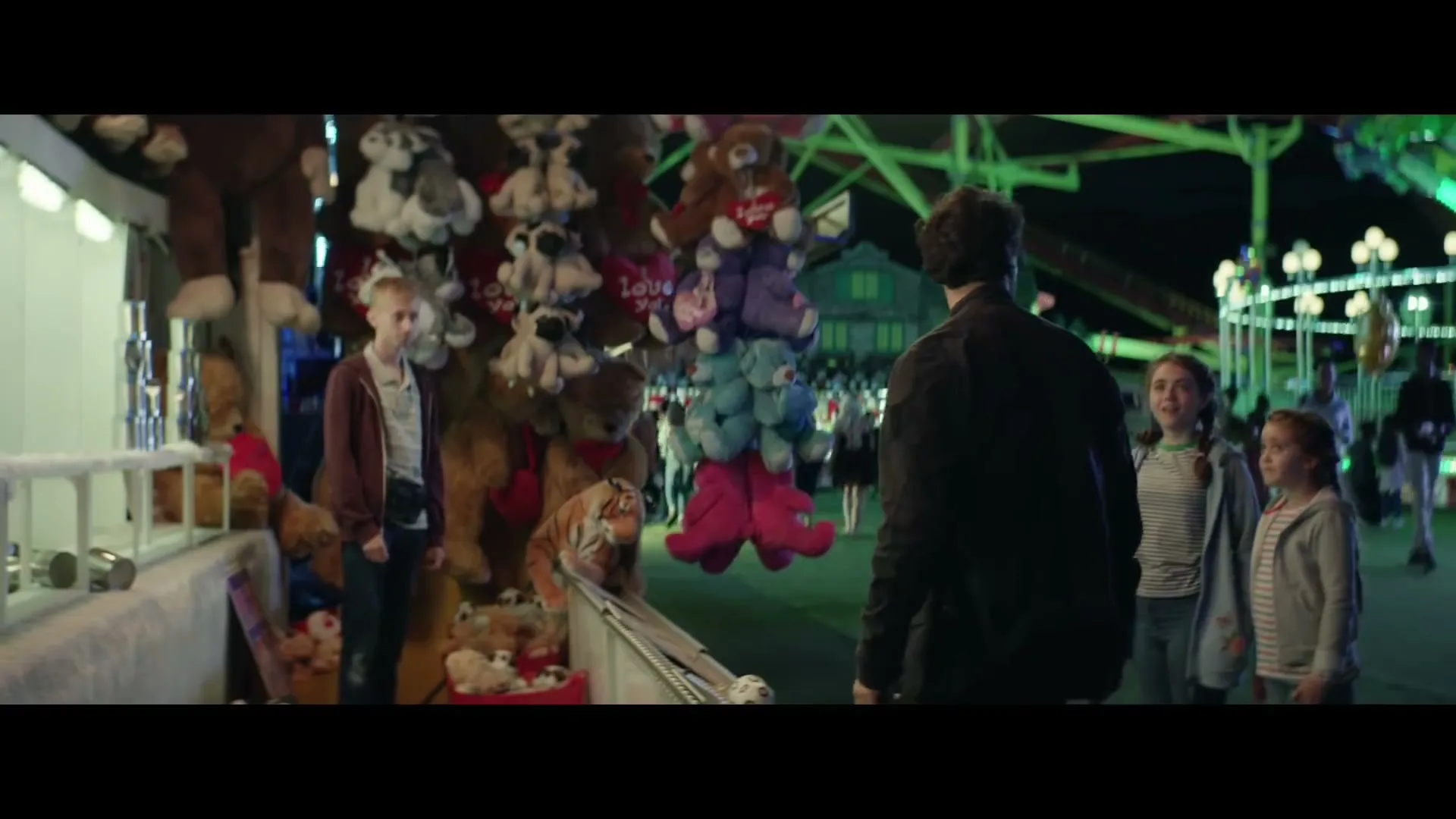 A man with dark hair and a beard stands with two young girls at a carnival game booth filled with stuffed animals at night.
