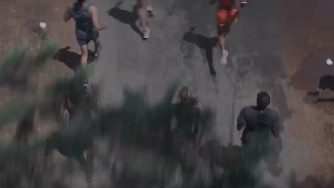 An overhead shot shows a diverse group of people running on a dirt path, surrounded by green foliage, during the day.