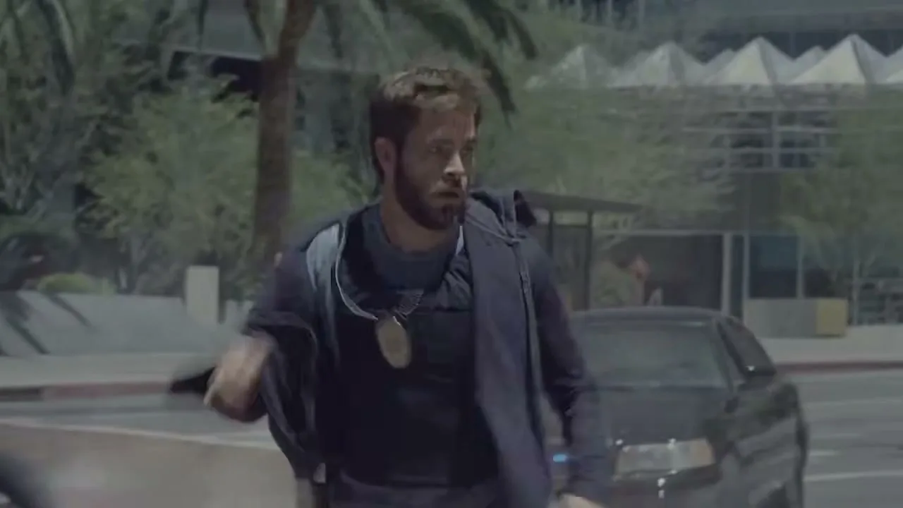 A male police officer with a beard and dark uniform runs across a city street during the day, with buildings and palm trees in the background.
