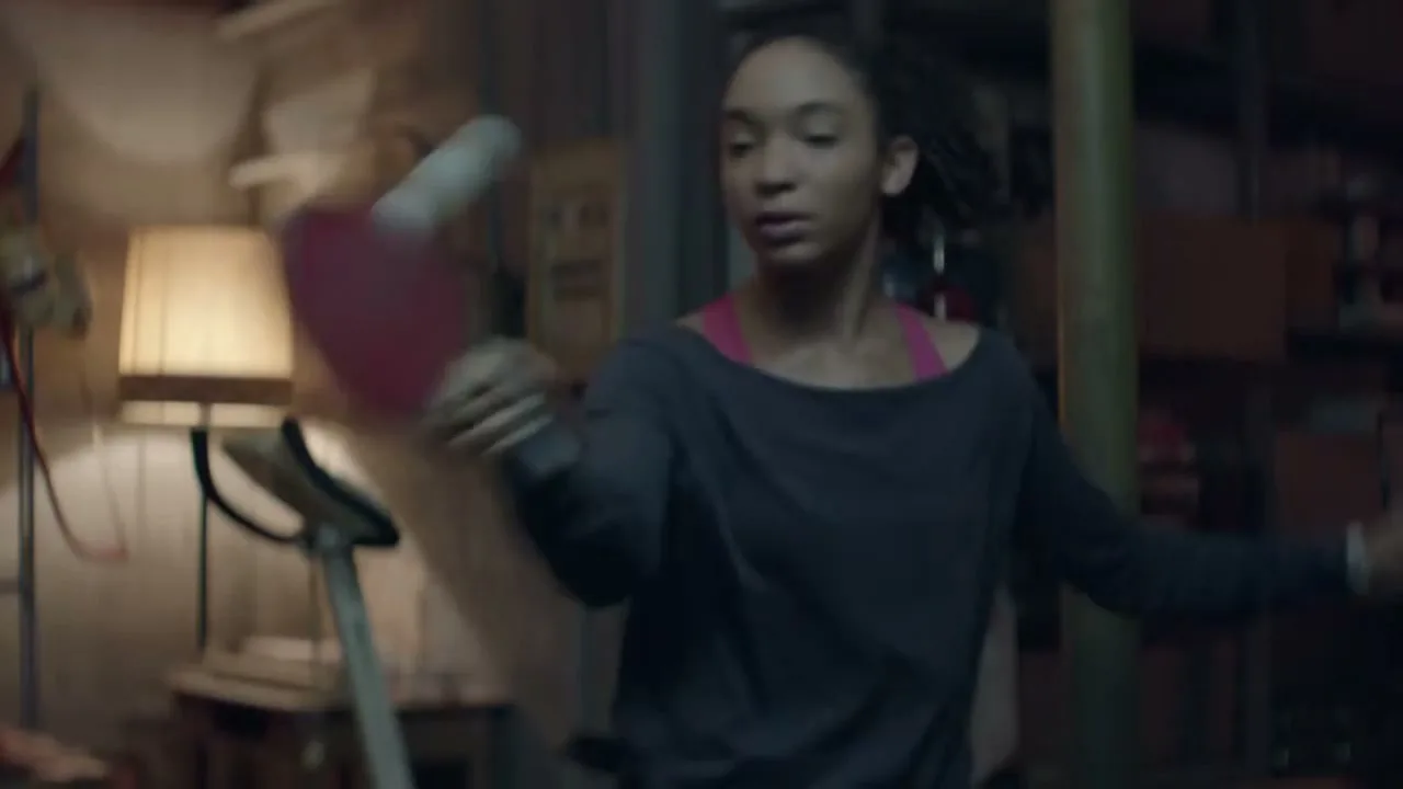 A young woman with dark hair practices table tennis alone in a dimly lit, cluttered basement, hitting a ball against a wall.