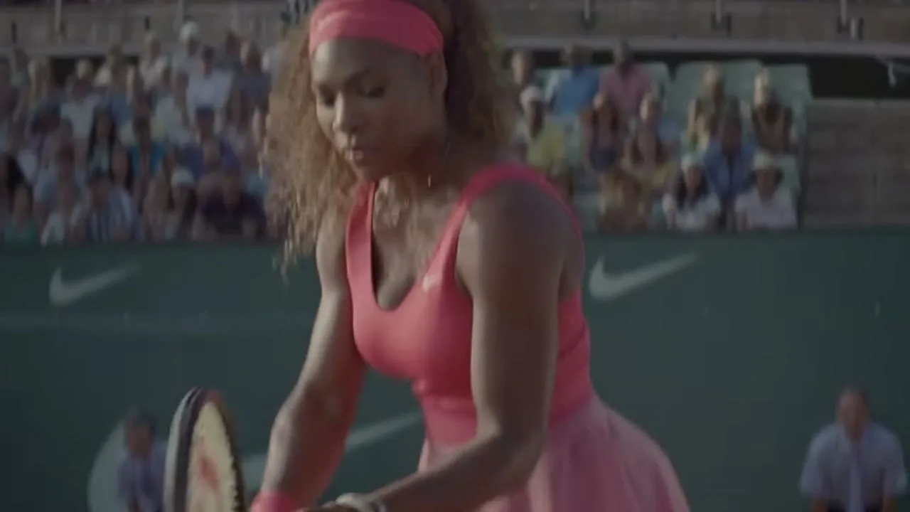 Serena Williams, a female tennis player in a pink dress, prepares to serve on a clay court during a match, with spectators in the background.