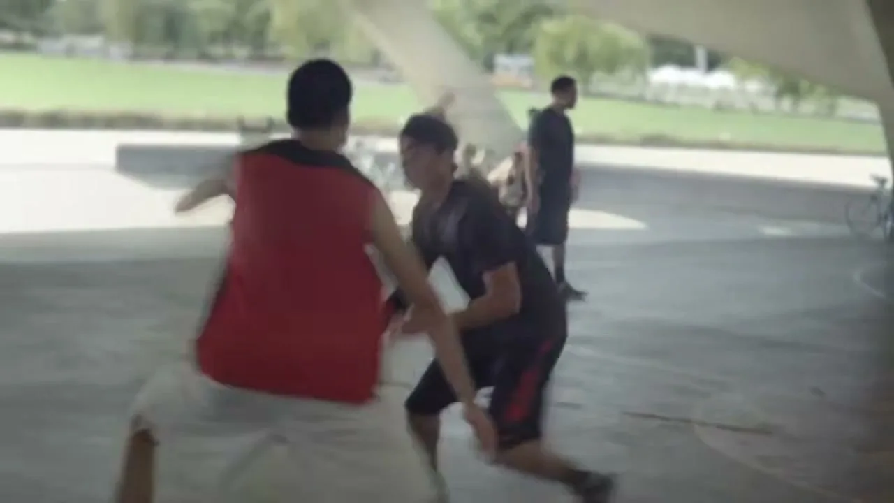 Two young men play basketball on an outdoor court under a bridge, with other people in the background.