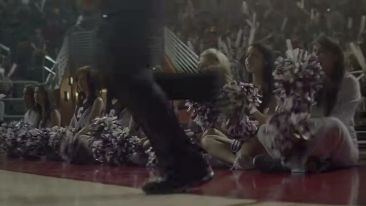 A group of female cheerleaders in dark uniforms with purple and white pom-poms sit on a red basketball court, looking up at someone passing by.