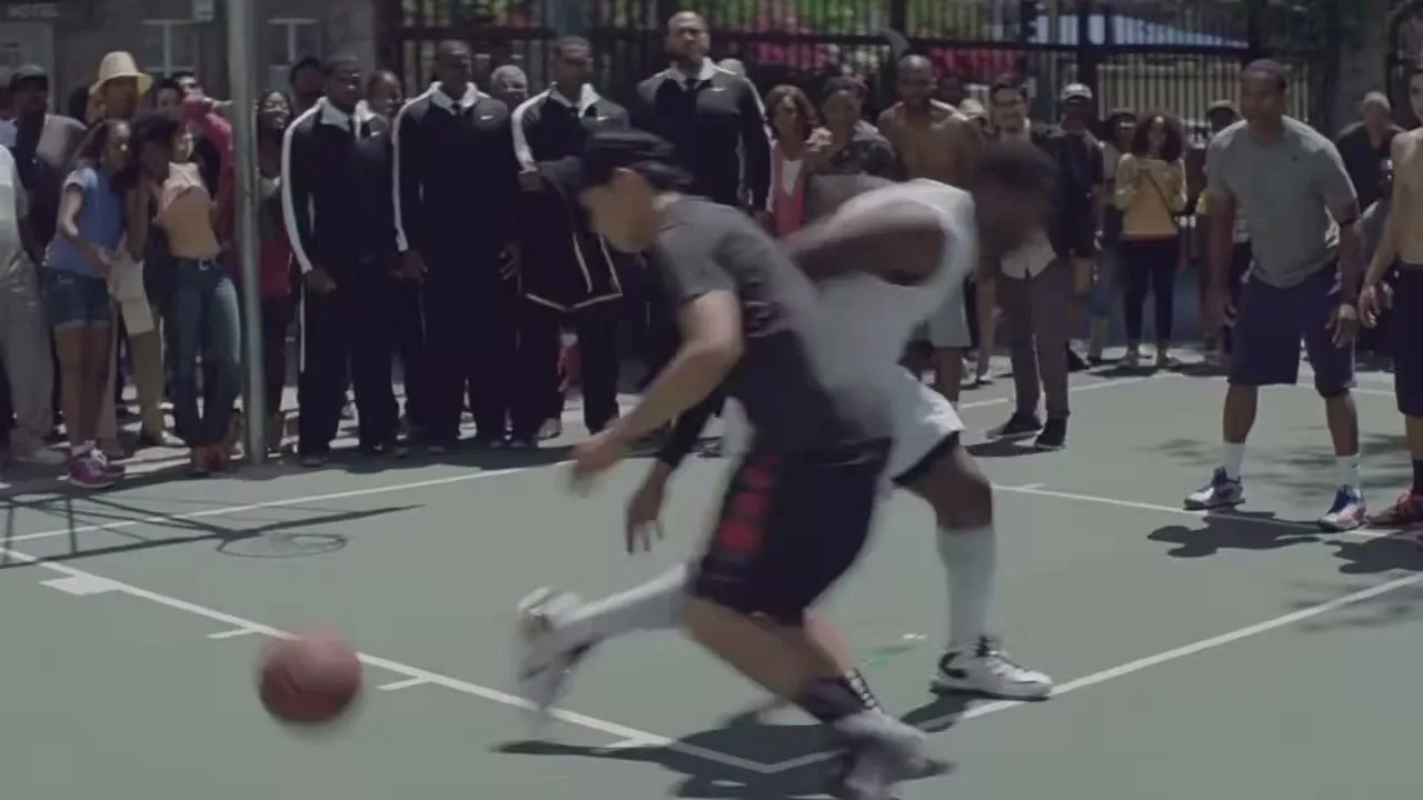 Two male basketball players dribble and defend each other on an outdoor court, surrounded by a crowd of spectators on a sunny day.