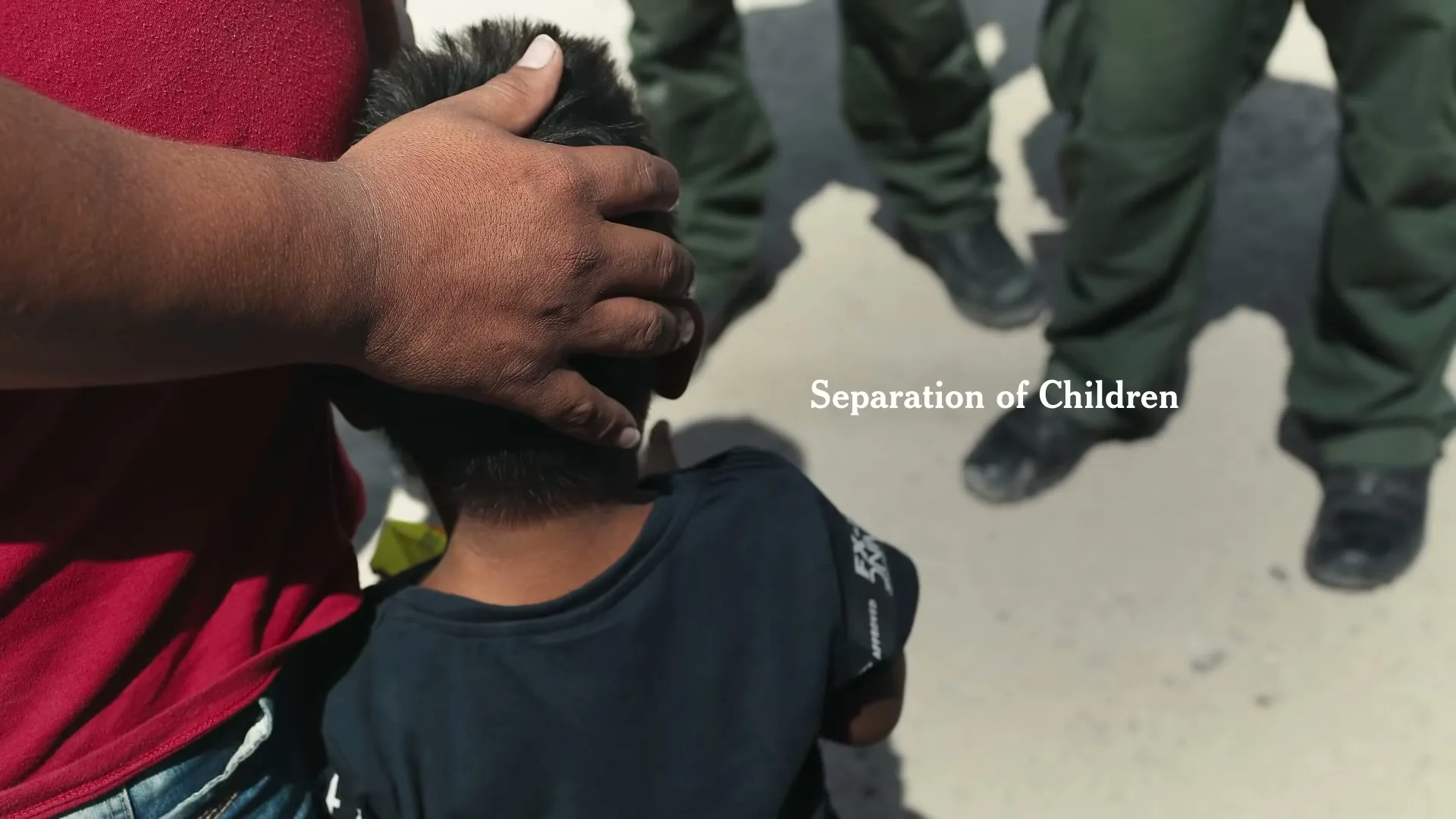 A man in a red shirt places his hand on a child's head, with two pairs of uniformed legs visible in the background, on a sunny day outdoors.