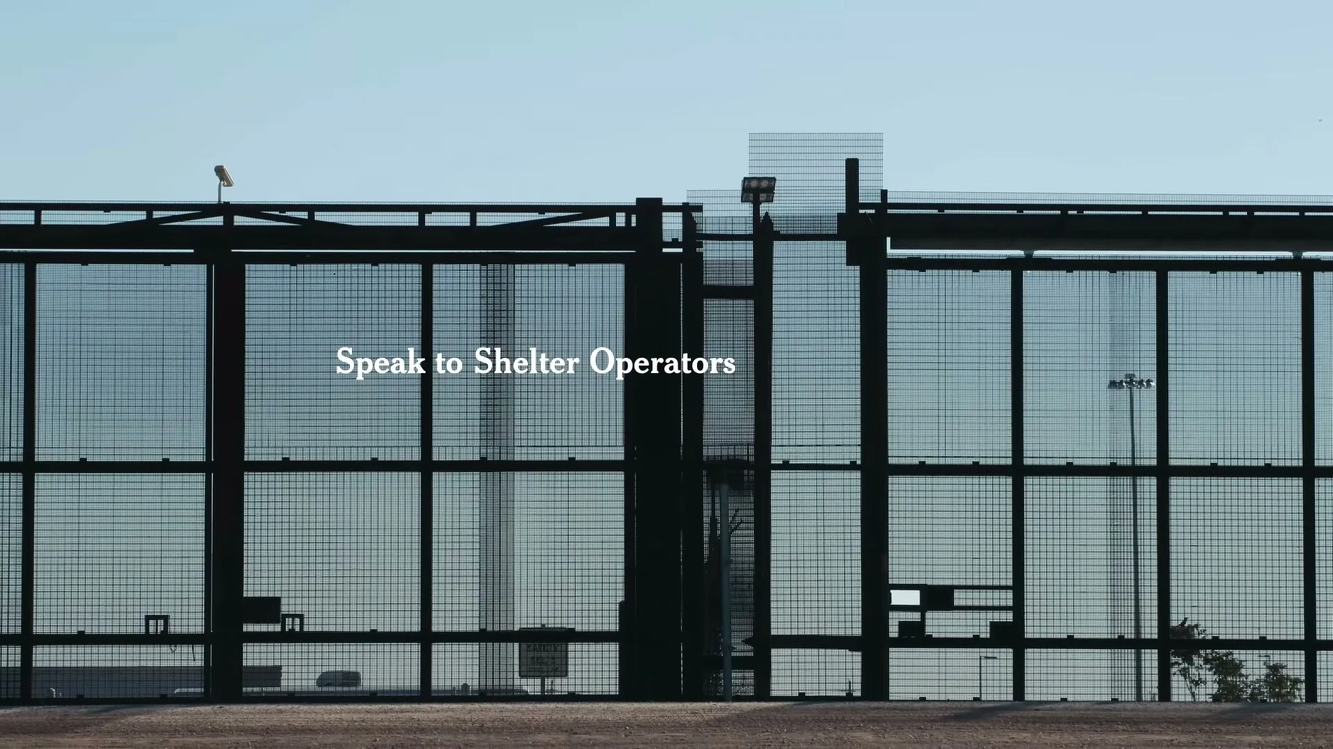 A wide shot of a tall, dark metal fence with a clear blue sky in the background, with the text "Speak to Shelter Operators" overlaid.