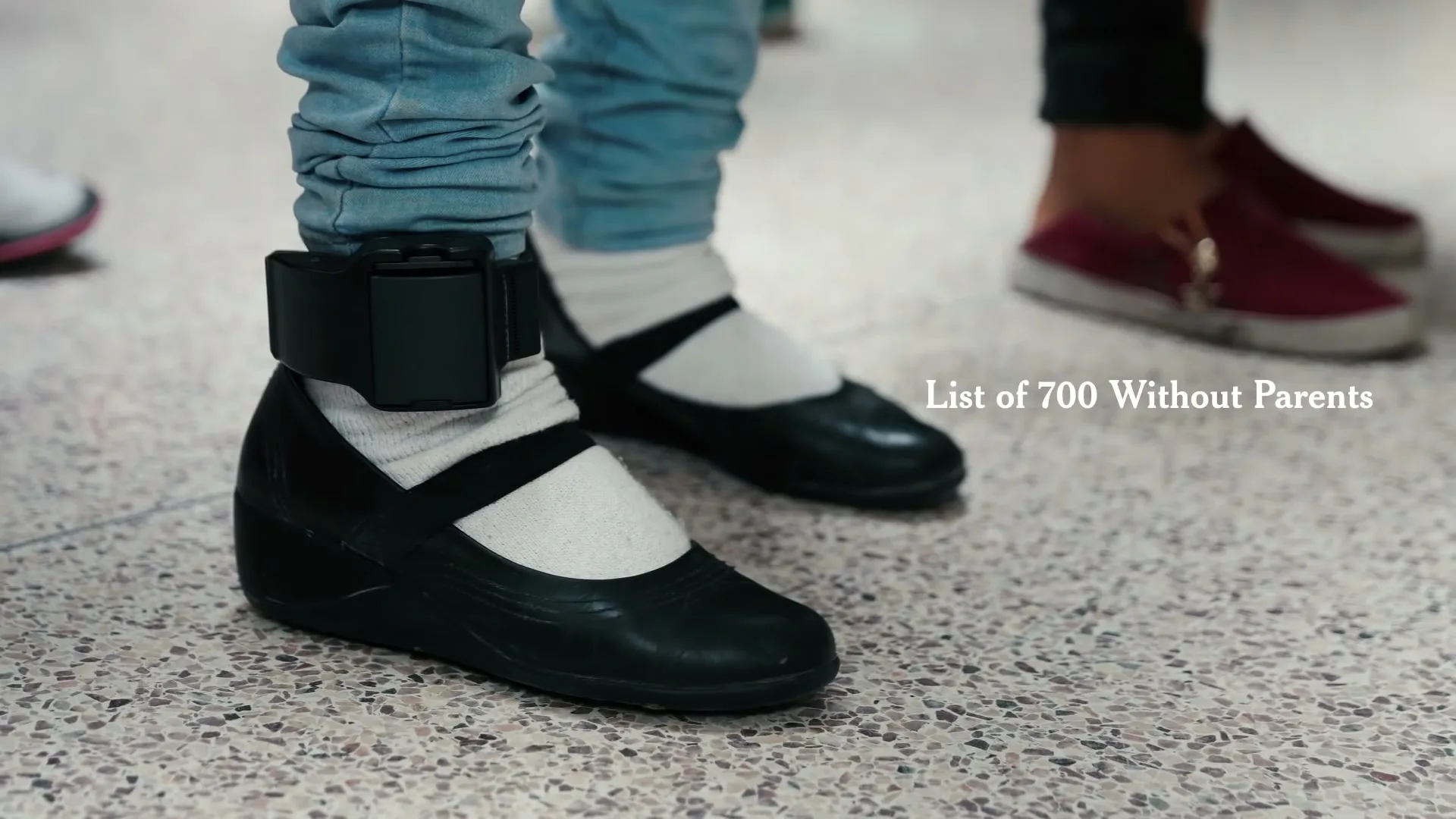 A child's leg with an ankle monitor is visible, wearing light blue jeans and black shoes, standing on a speckled floor with other people's feet in the background.