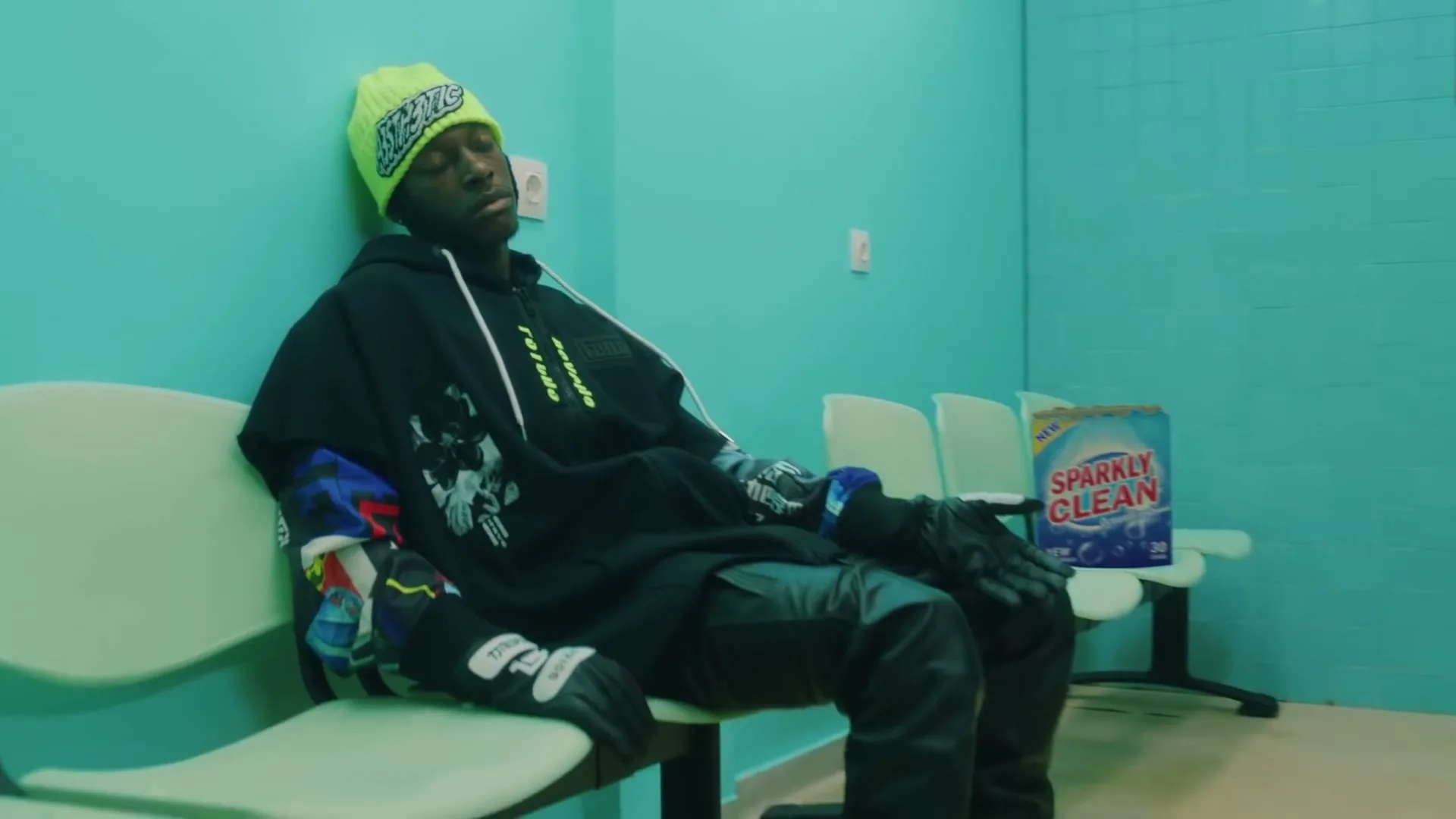 A young Black man in a neon green beanie and black hoodie sits on a bench in a turquoise laundromat, looking bored.