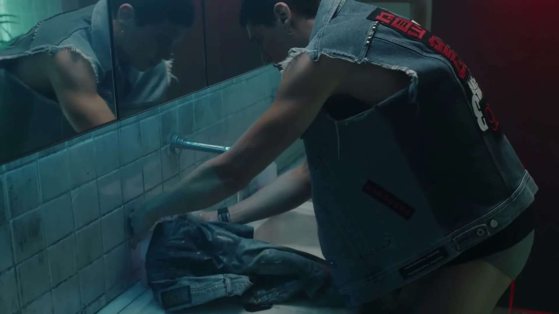 A young man in a denim vest washes a pair of jeans in a public restroom sink, with his reflection visible in the mirror.