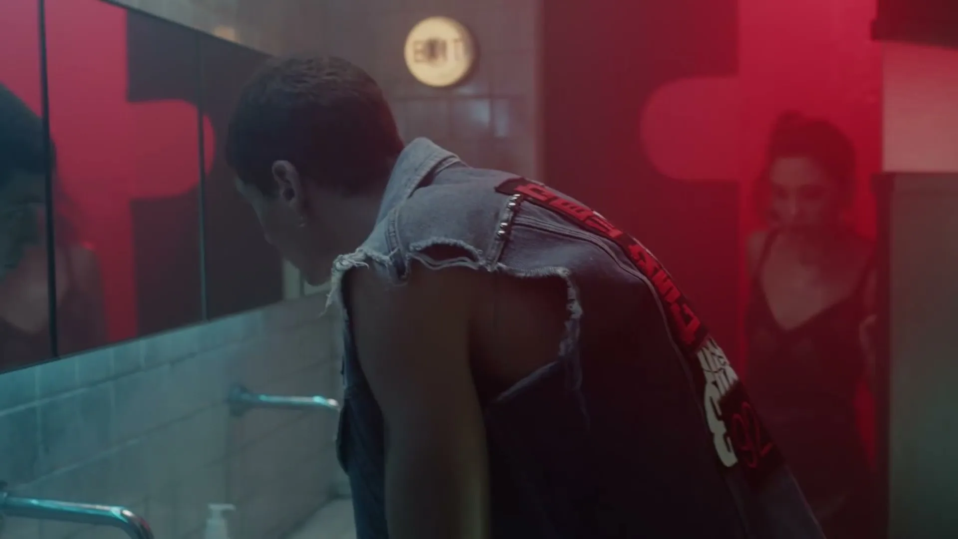 A young man in a ripped denim vest washes his face in a dimly lit public restroom, as a young woman with dark hair approaches him from behind.