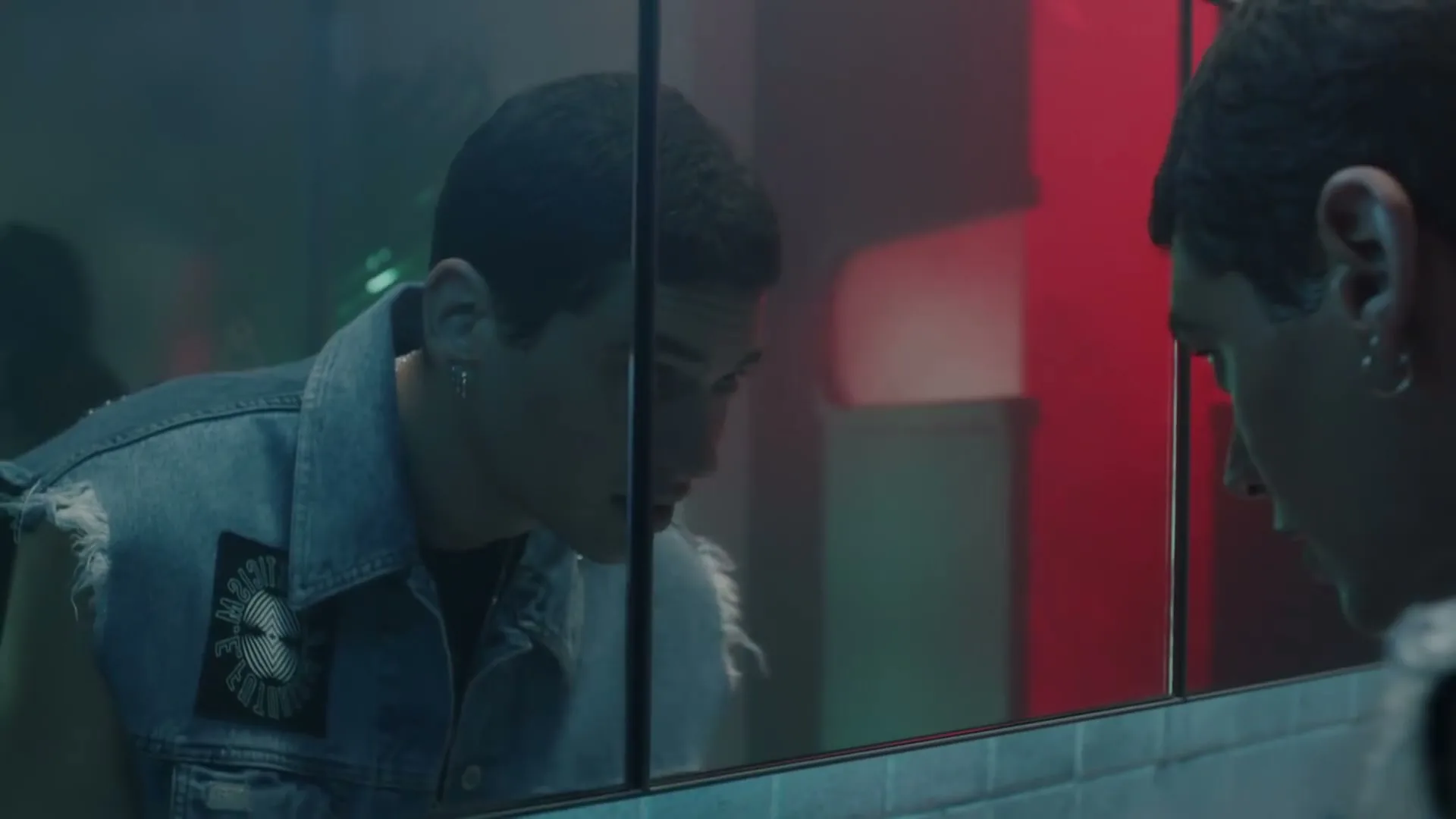 A young man with short dark hair and a denim vest looks at his reflection in a mirror in a dimly lit, red-hued bathroom.