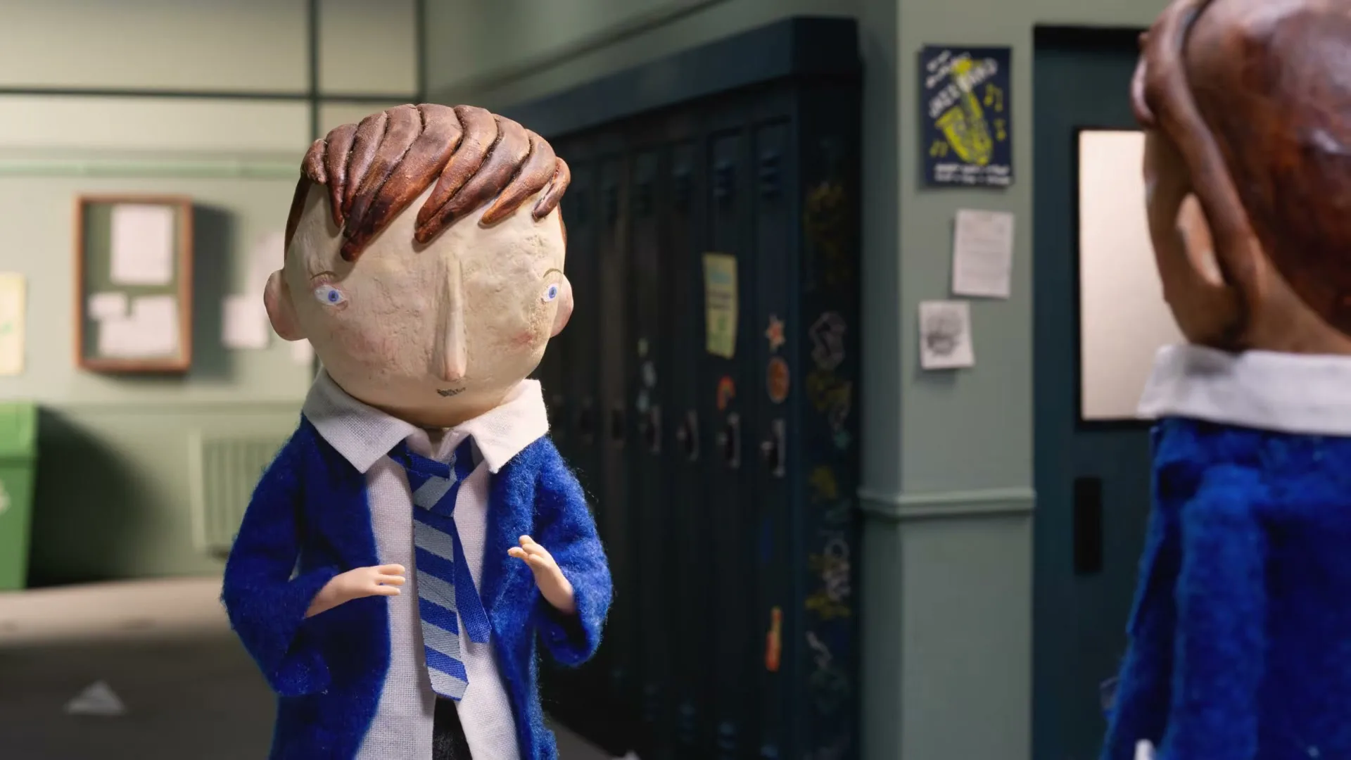 A stop-motion animated boy in a blue school uniform and striped tie looks at another boy in a school hallway.
