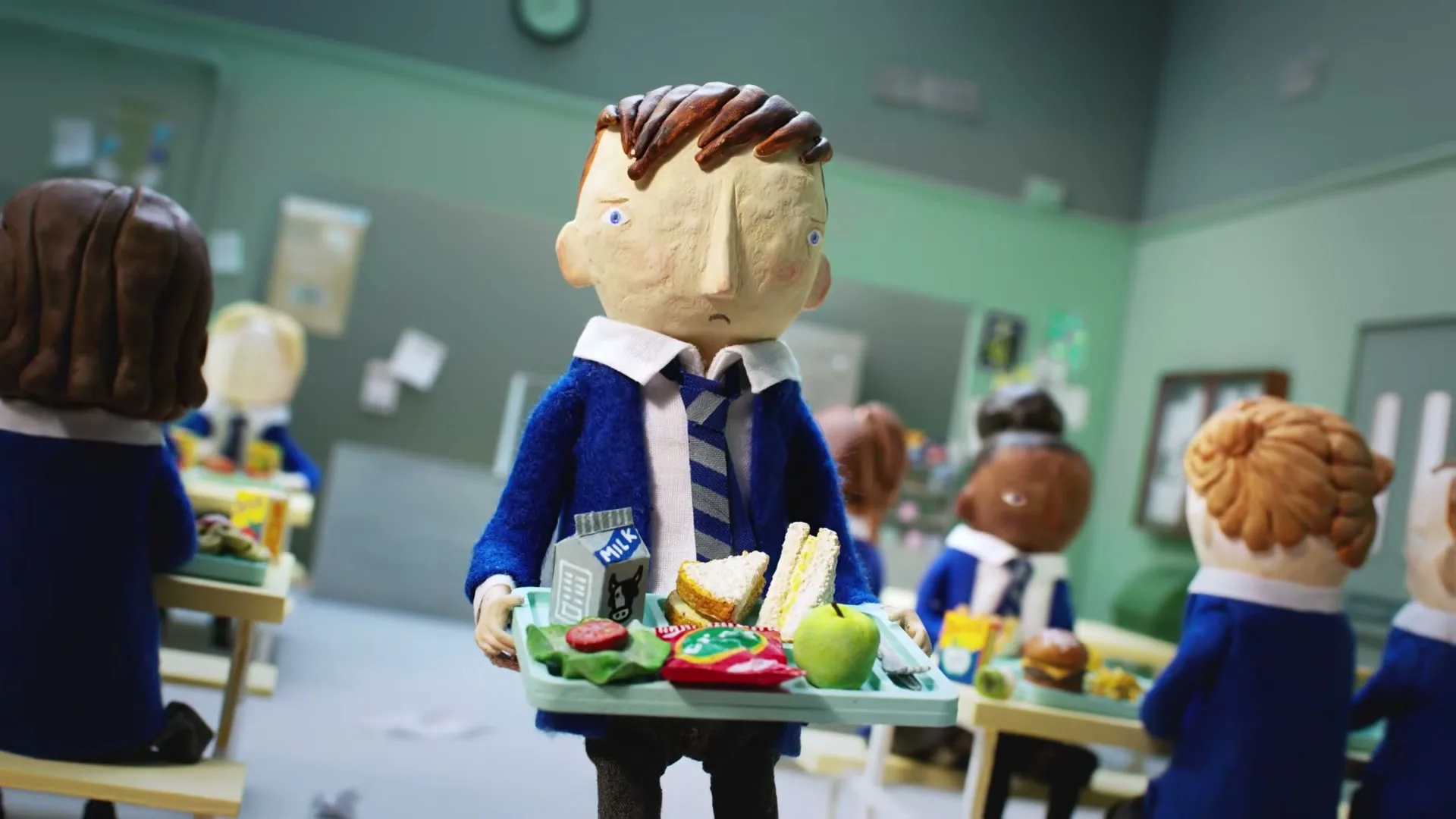 A stop-motion animated boy in a school uniform holds a lunch tray, looking sad in a busy school cafeteria.