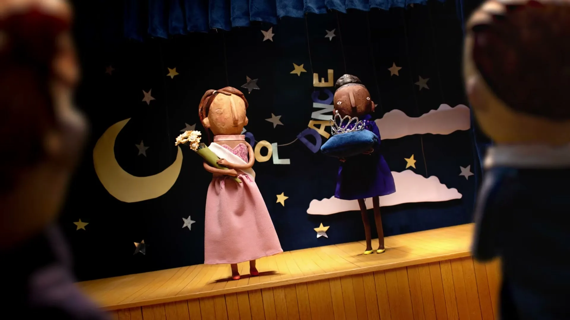 Two stop-motion animated female figures stand on a stage, one in a pink dress holding flowers, the other in a purple dress holding a crown.