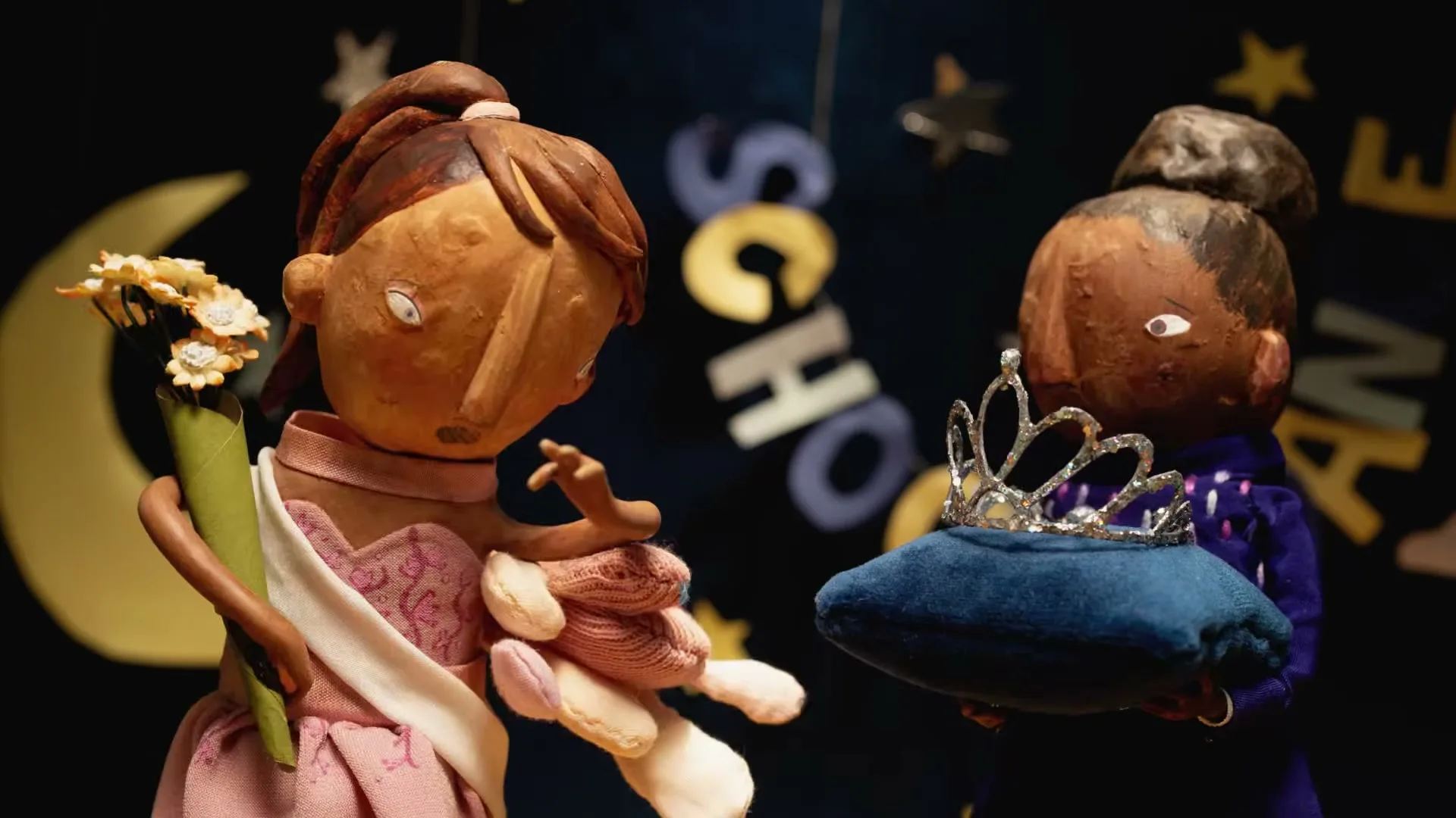 Two stop-motion puppets, one wearing a sash and holding flowers, the other holding a crown on a pillow, stand against a dark background with hanging letters.