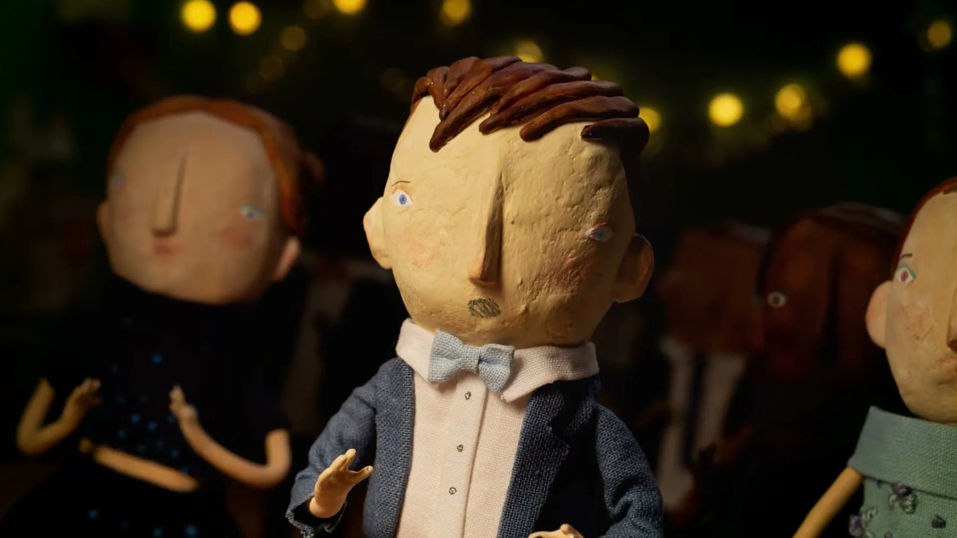 A stop-motion puppet of a young male in a suit and bow tie stands among other puppets, looking around with a neutral expression in a dimly lit room.