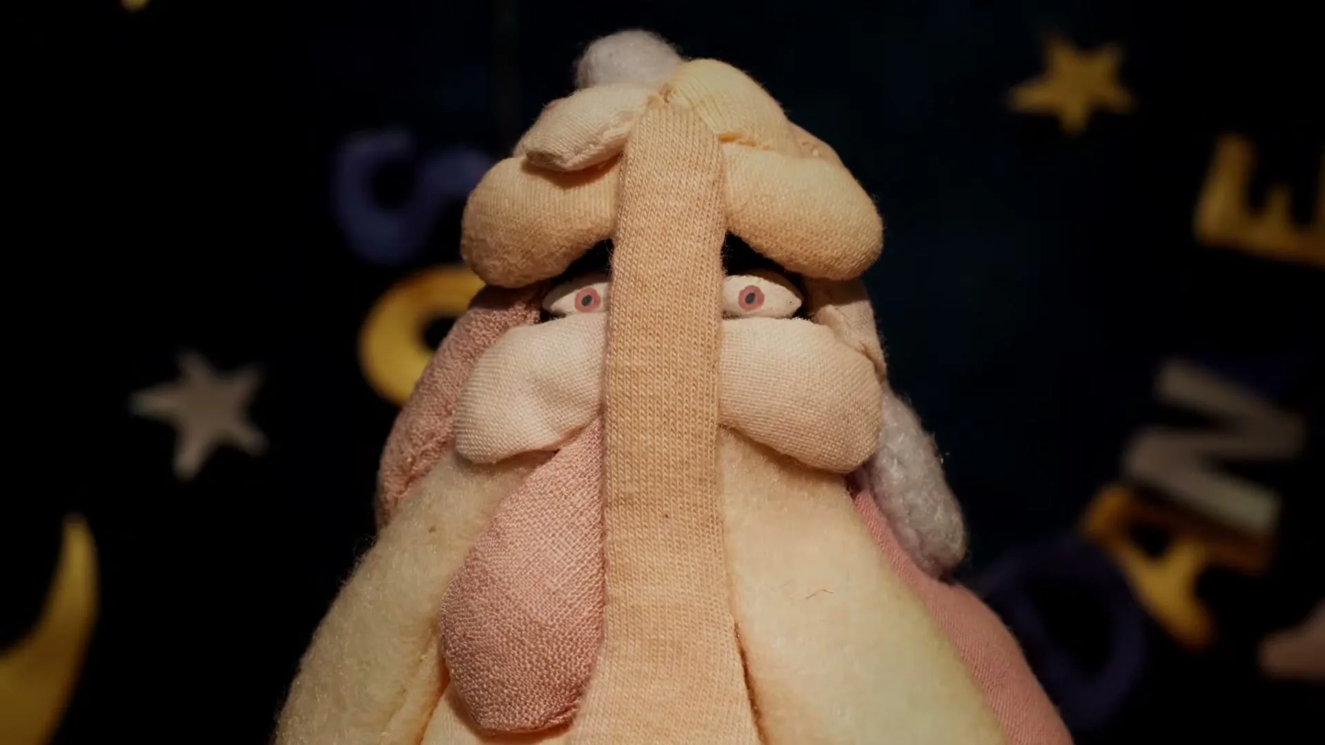 A handmade, fabric puppet with large, wide eyes and a long, flat nose looks directly at the camera against a dark background with blurry shapes.