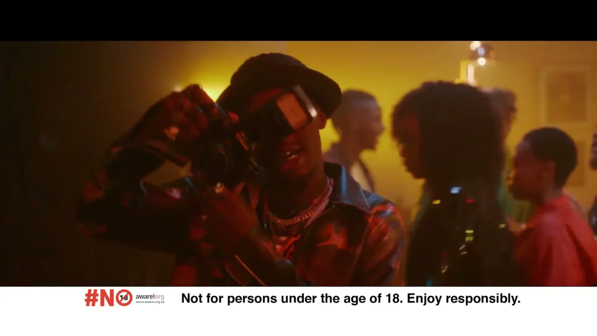 A young man in a hat and sunglasses holds a camera with a flash, smiling at a party with other people in the background.