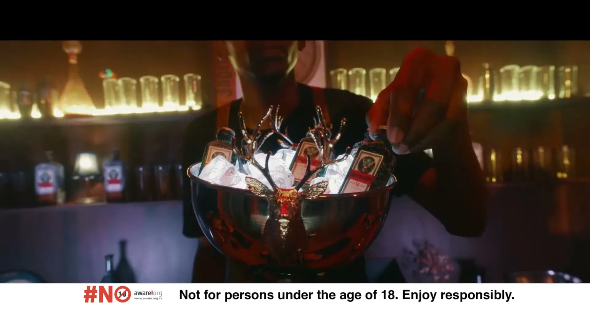 A bartender in a dark bar holds a Jägermeister bottle over a silver bowl filled with ice and mini Jägermeister bottles.