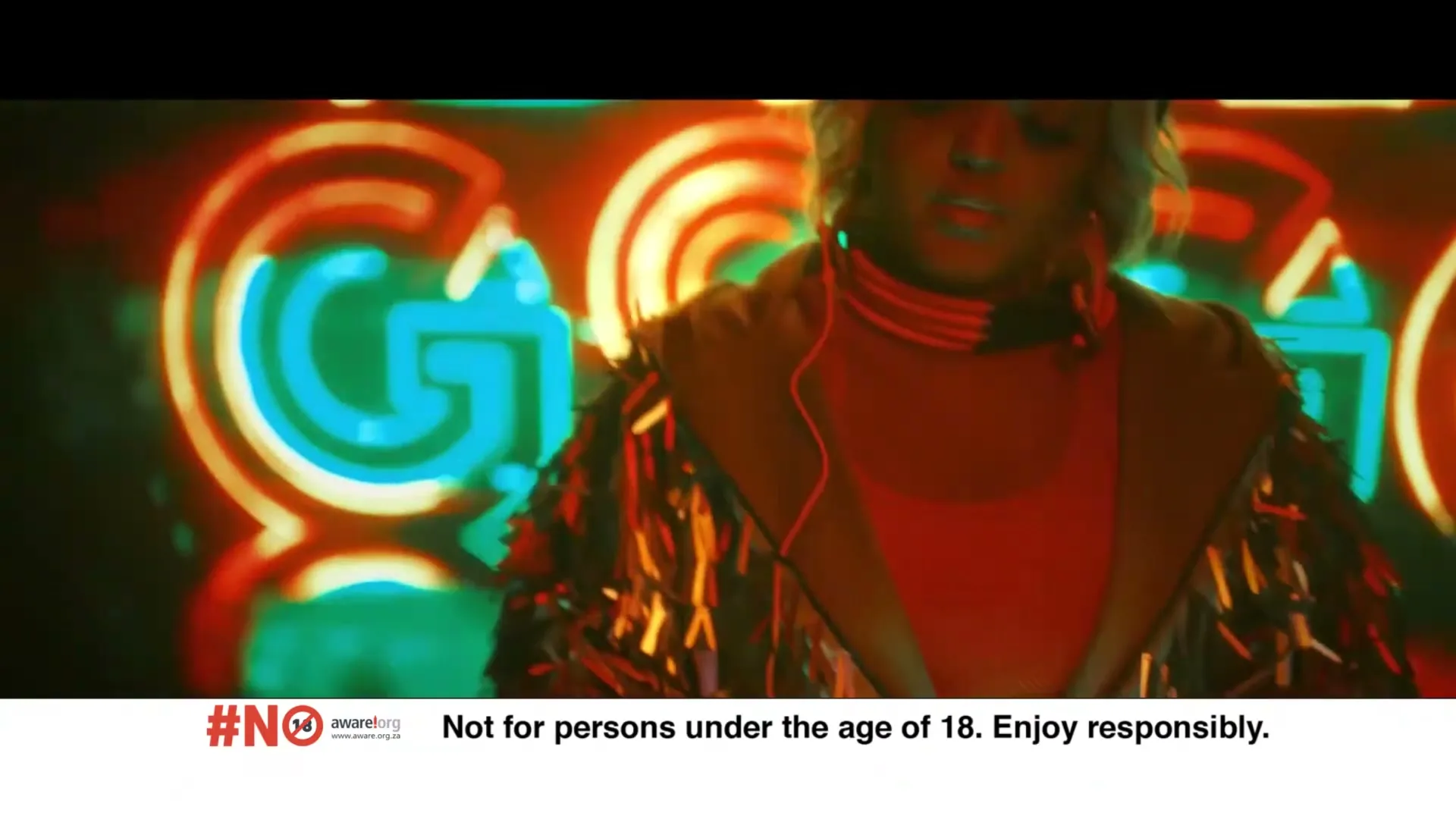 A person with blonde hair and a shimmering jacket stands in front of neon lights, looking down with a serious expression.