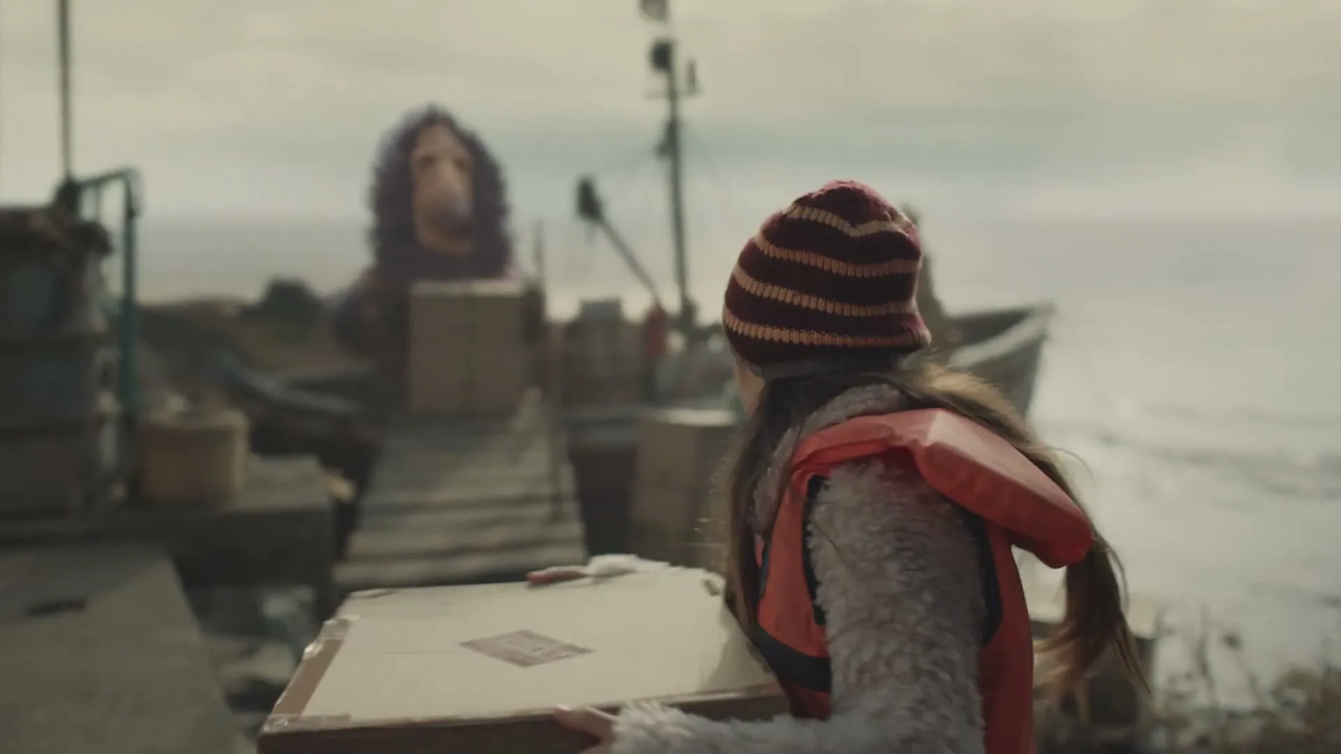 A young girl in a beanie and life vest carries a box towards a boat on a dock, where two large, furry creatures are waiting.